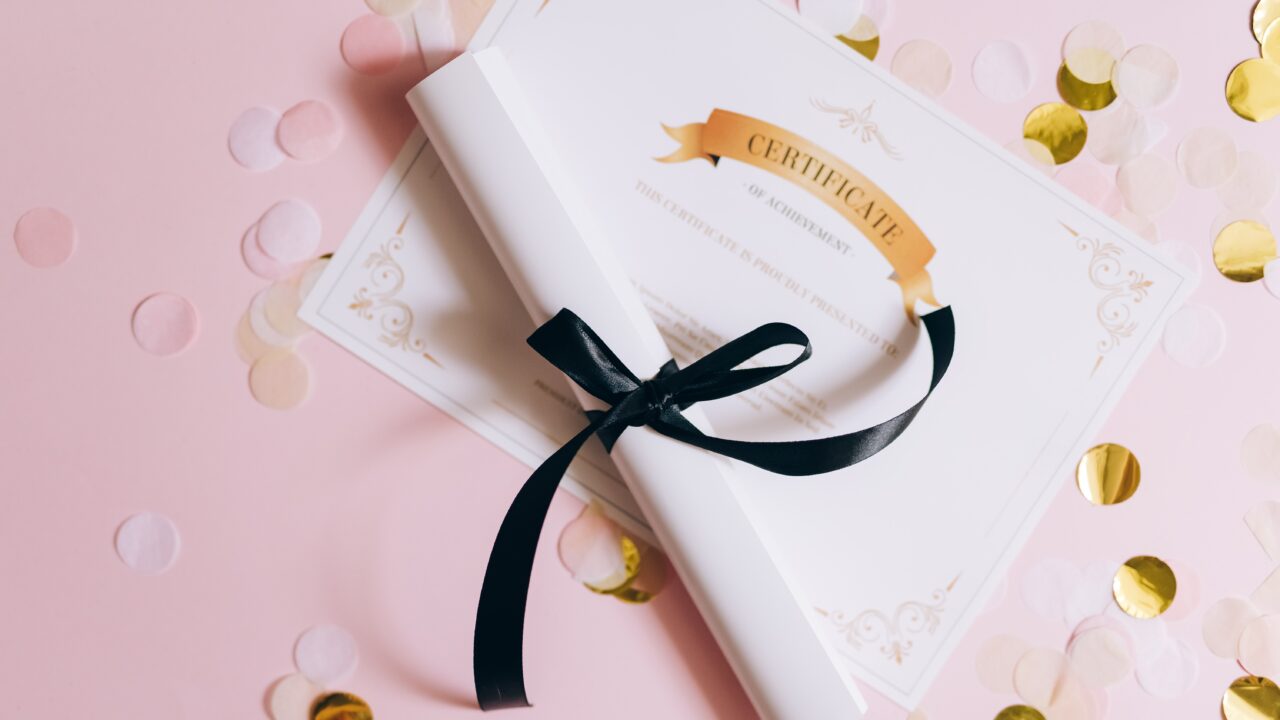 One flat membership certificate and one rolled up and tied with ribbon, both laid on gold confetti.